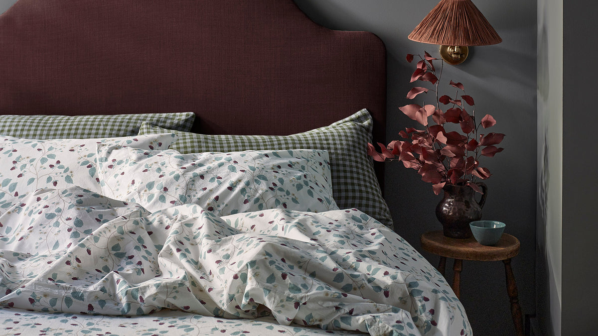 A styled bedroom scene showing a bed with white bedding printed with small blue and burgundy floral motifs, green-and-white checkered pillows, a dark burgundy upholstered headboard, a bedside table with a lamp, and dried burgundy foliage against a dark gray wall.