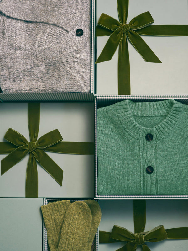 A gift box arrangement featuring folded knit garments and ribbons. Visible items include a light gray knit top, a sage green knit cardigan with dark buttons, and olive green ribbed knit fabric, each styled with an olive green ribbon bow on a pale background.