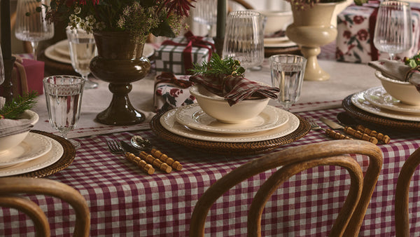 A styled holiday table setting with a red and white checkered tablecloth, white ceramic plates, gold-rimmed placemats, glass water cups, and a rustic floral centerpiece.