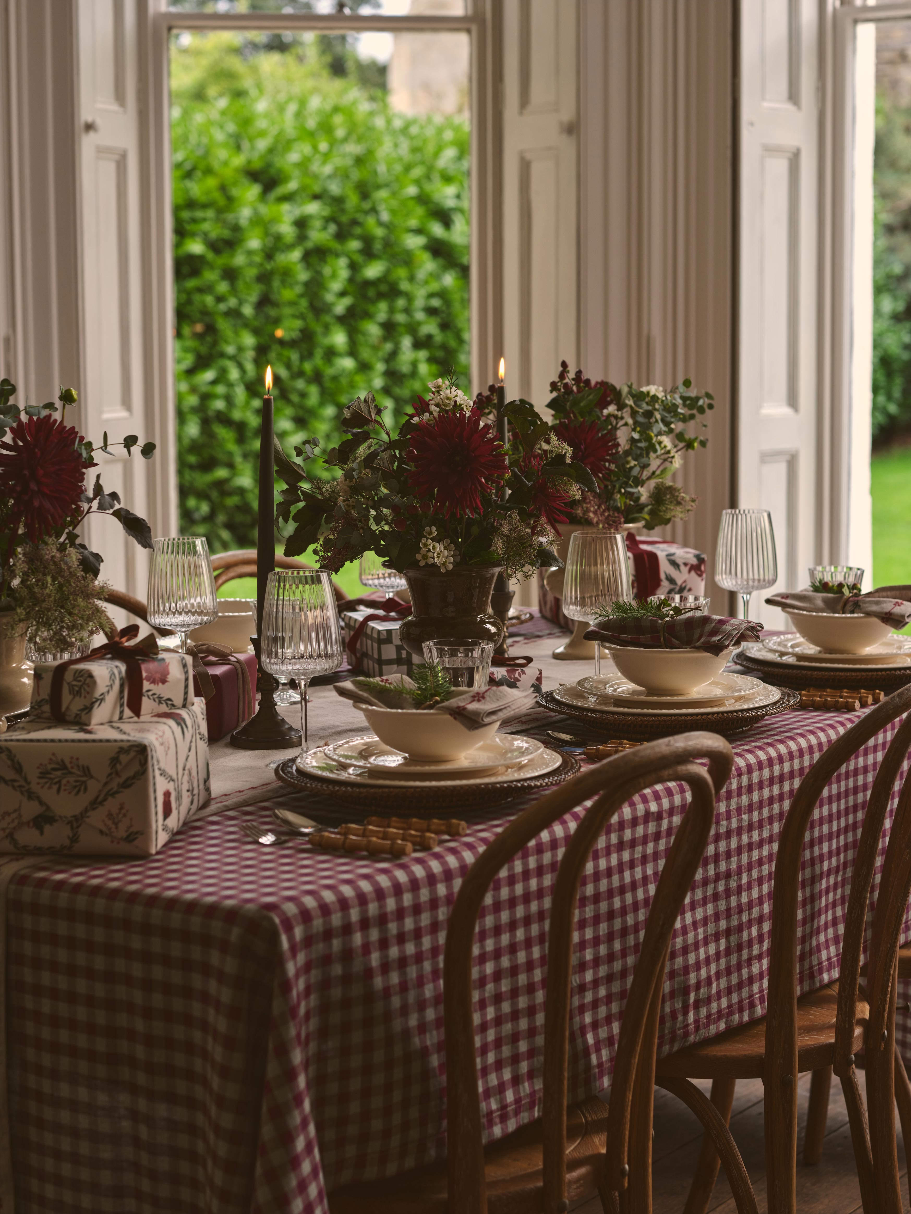 A styled dining table set with a red and white gingham tablecloth, white plates, dark red floral centerpieces, lit taper candles, and wooden chairs in a bright room with large windows overlooking greenery.
