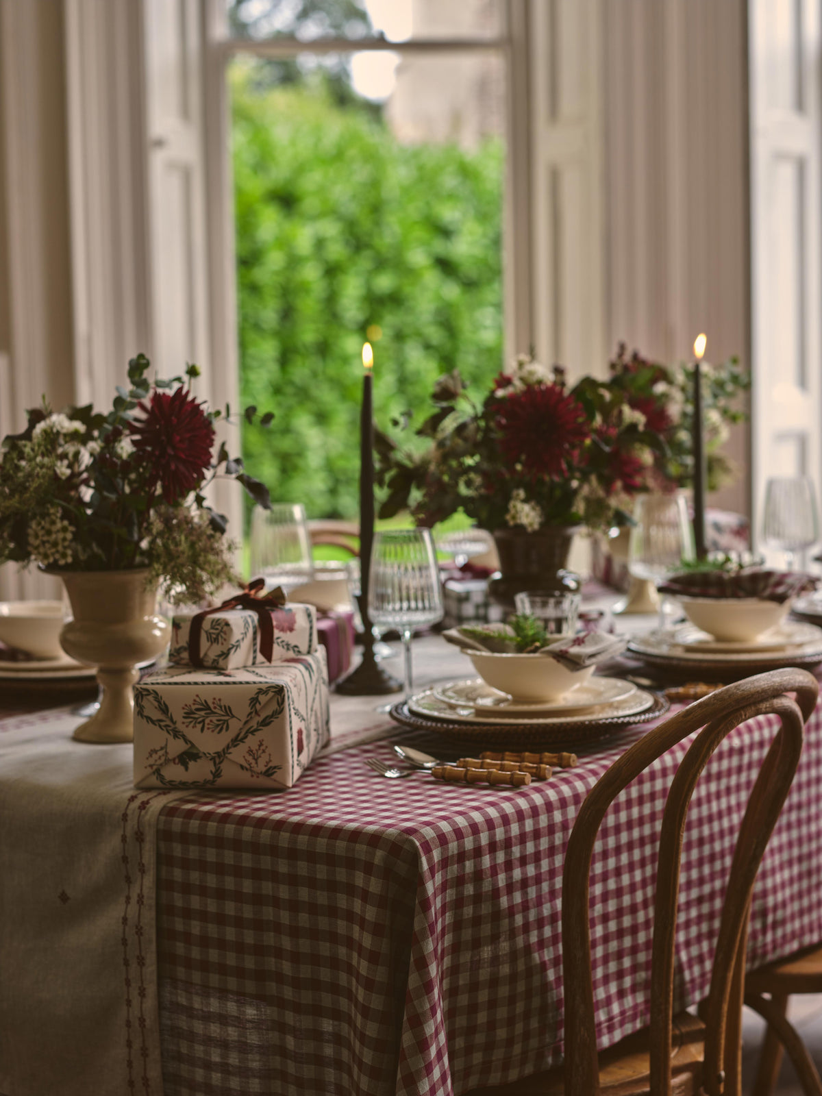 A styled dining table set with a red and white gingham tablecloth, white plates, black taper candles, floral centerpieces, and wrapped gift boxes in a softly lit indoor setting near a window.