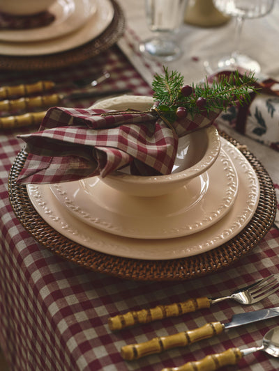 A styled table setting featuring a cream-colored ceramic bowl and matching plates layered over a brown woven charger plate, set on a red and white checkered tablecloth. A matching checkered napkin is folded inside the bowl and decorated with a sprig of green pine and red berries.