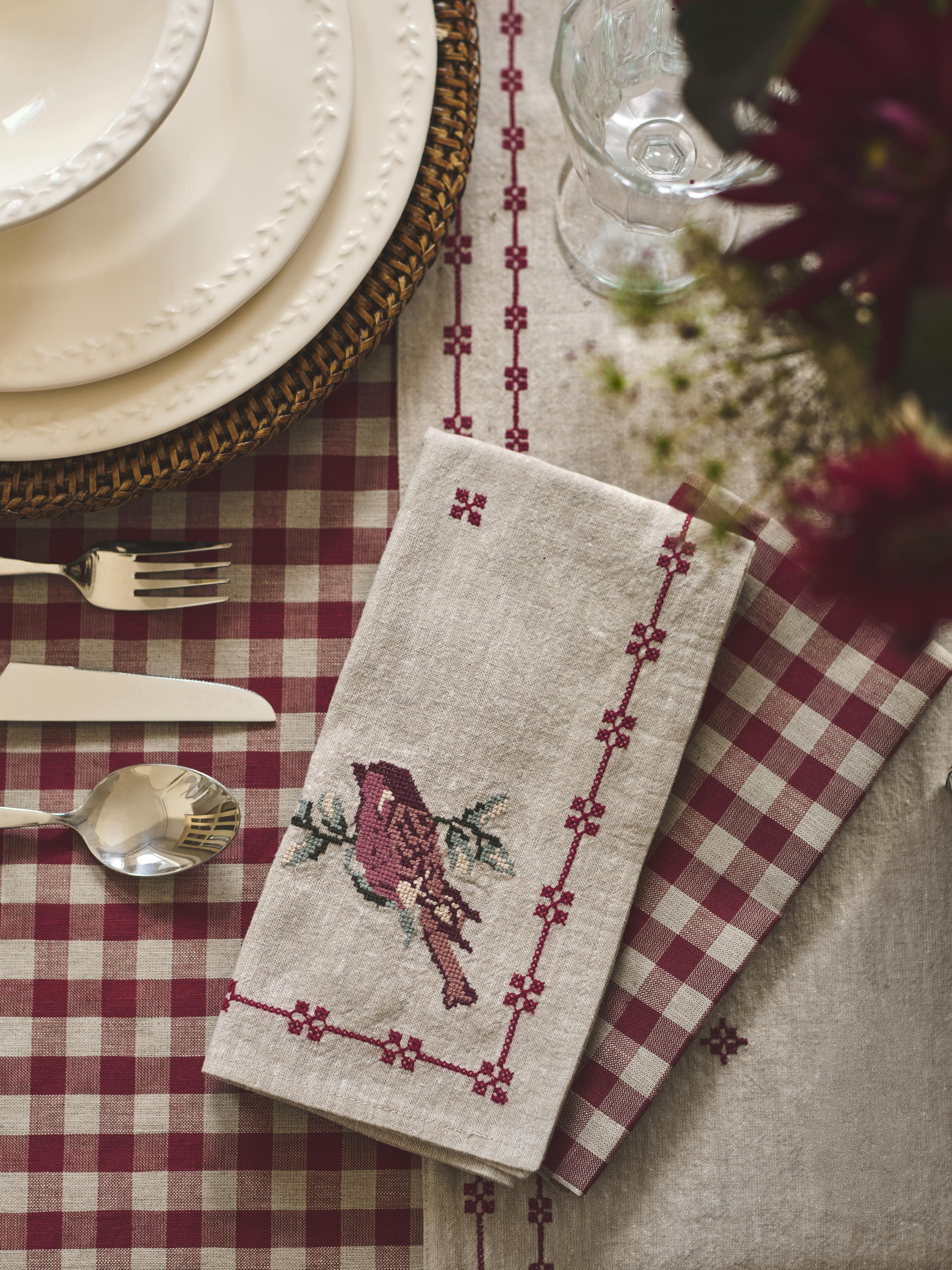 A folded linen napkin with a red bird and floral border embroidery, styled on a red and white checkered tablecloth with plates, cutlery, and a glass.