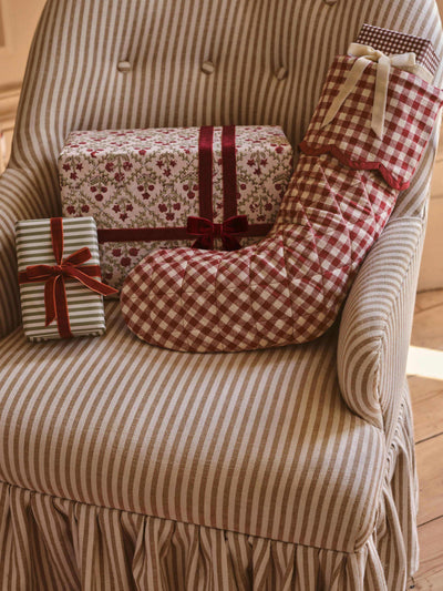 A cozy holiday-themed product display featuring a red and white gingham Christmas stocking, a matching red and white gingham gift bag, and two wrapped presents on a beige and brown striped armchair.