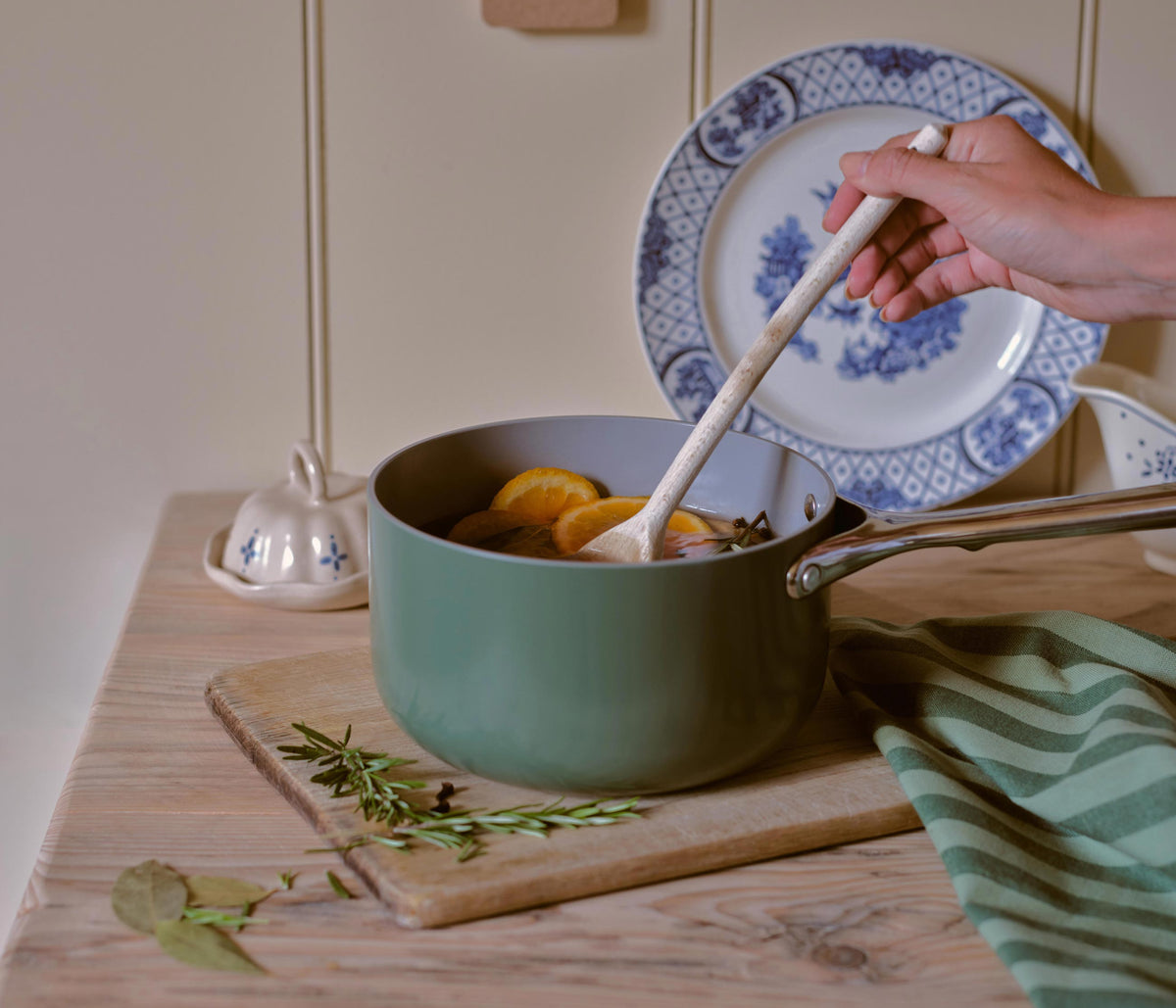 A sage green ceramic saucepan with a wooden spoon is shown on a wooden cutting board, containing citrus slices. A hand holds the spoon, and a blue and white patterned plate is propped behind the pot on a cream-colored wall.