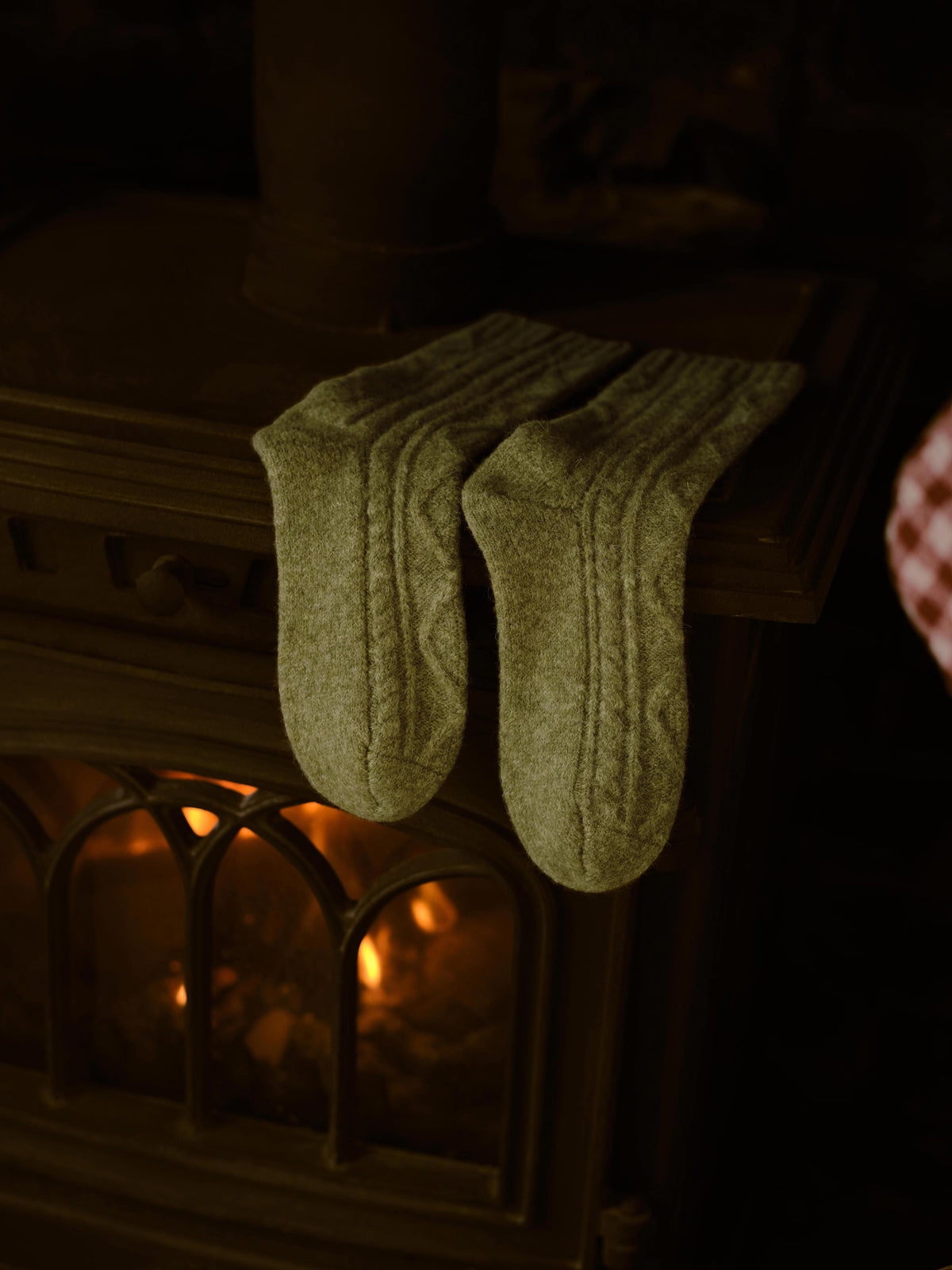A pair of chunky cable-knit socks in a warm olive green color, draped over the edge of a vintage wood-burning stove with a glowing fire inside.