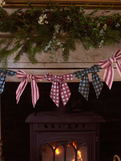 A rustic holiday garland draped over a dark fireplace opening, featuring a large pink striped bow in the center, a muted green gingham bow on the left, and a burgundy gingham bow on the right, with pine branches and small white berries above.