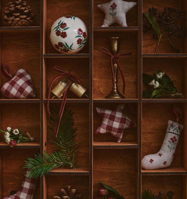 A rustic wooden shelf display filled with Christmas decorations, including a white floral ornament, red-and-white gingham stockings, pinecones, green pine branches, a brass candle holder, and dried botanicals.