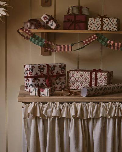 A styled holiday gift-wrapping display with multiple wrapped presents and wrapping paper on wooden shelves and a table, set against a beige paneled wall with a cream ruffled curtain below.