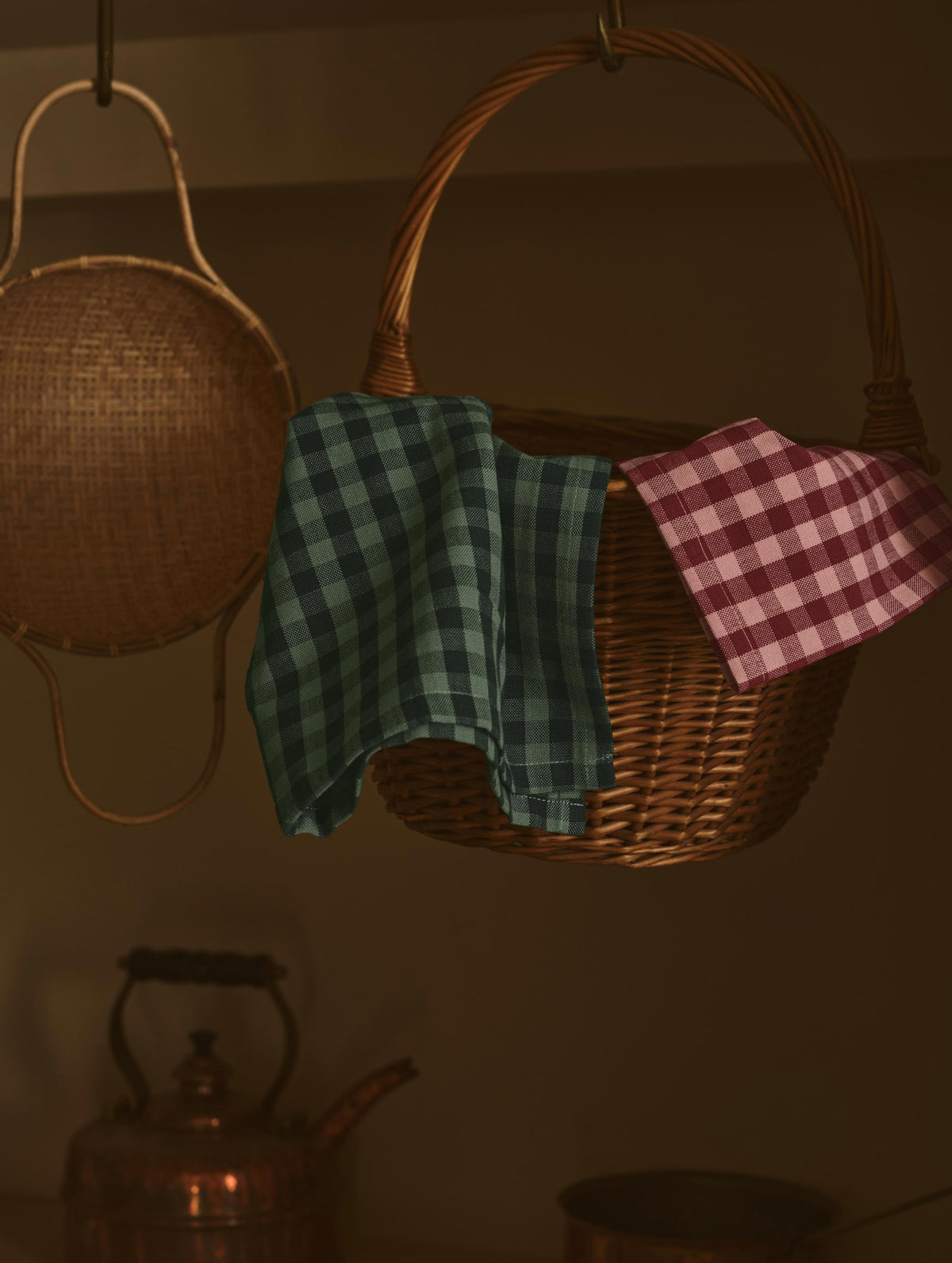 A rustic wicker basket hanging in a dimly lit interior, with two checkered cloth napkins draped over the sides—one green and blue gingham and one pink and red gingham. Another woven basket hangs to the left, and a copper kettle is visible in the background.