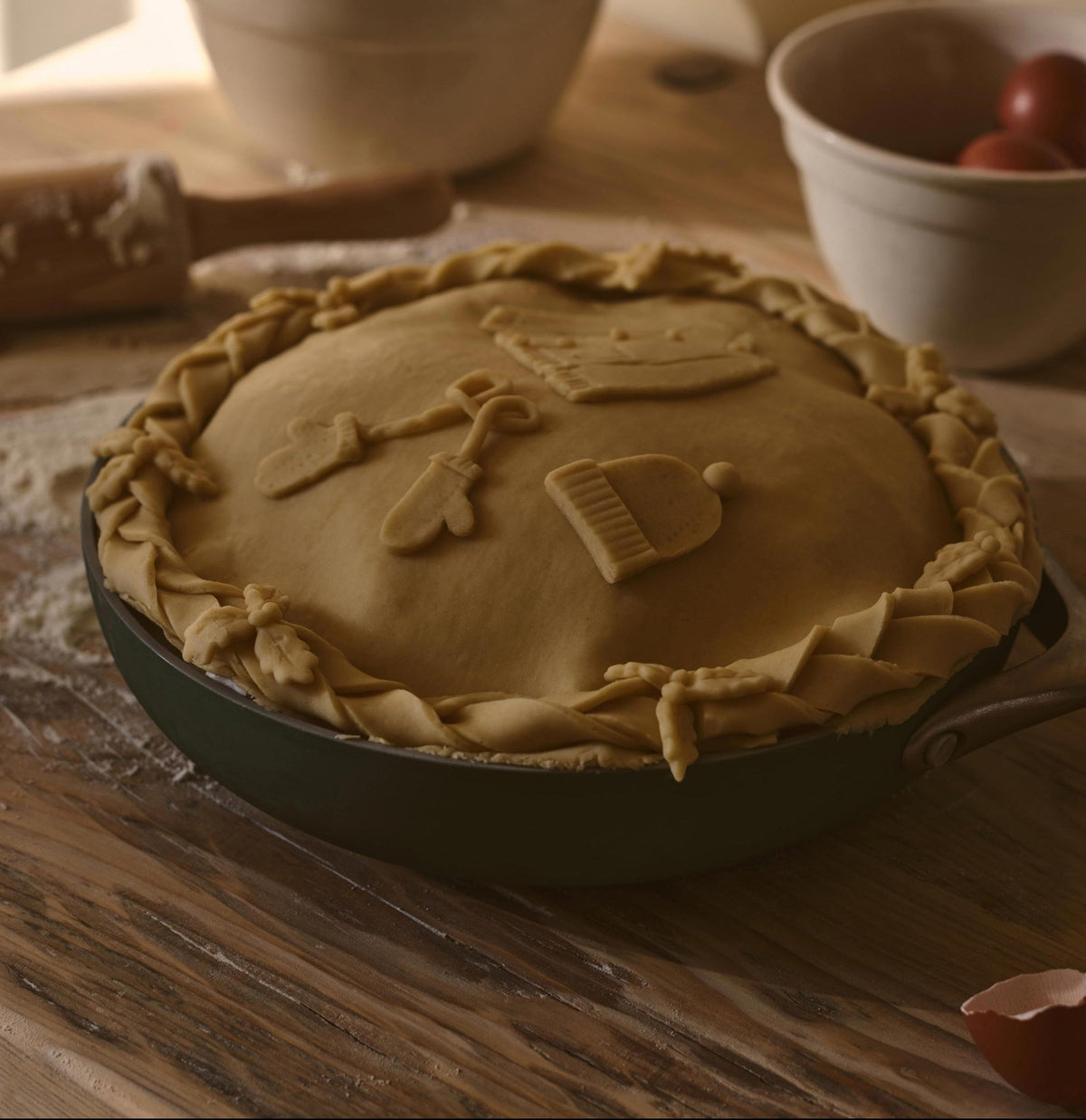 A baked pie in a dark cast iron skillet sits on a wooden table in a warm, rustic kitchen setting. The pie crust is decorated with small baked cookie shapes, and blurred bowls of ingredients are visible in the background.