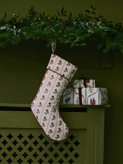 A Christmas stocking with a cream background and a red and green floral pattern hangs from a green garland above a mantel. Two wrapped gift boxes are stacked on the shelf to the right.