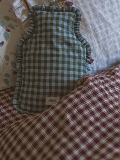 A green and white gingham check hot water bottle with ruffled edges, resting on a bed with a matching red and white checkered blanket.