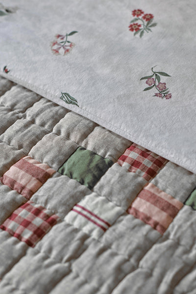 A close-up of a quilted patchwork blanket with a light gray base, featuring stitched floral motifs and gingham plaid accents in muted red, pink, and sage green.