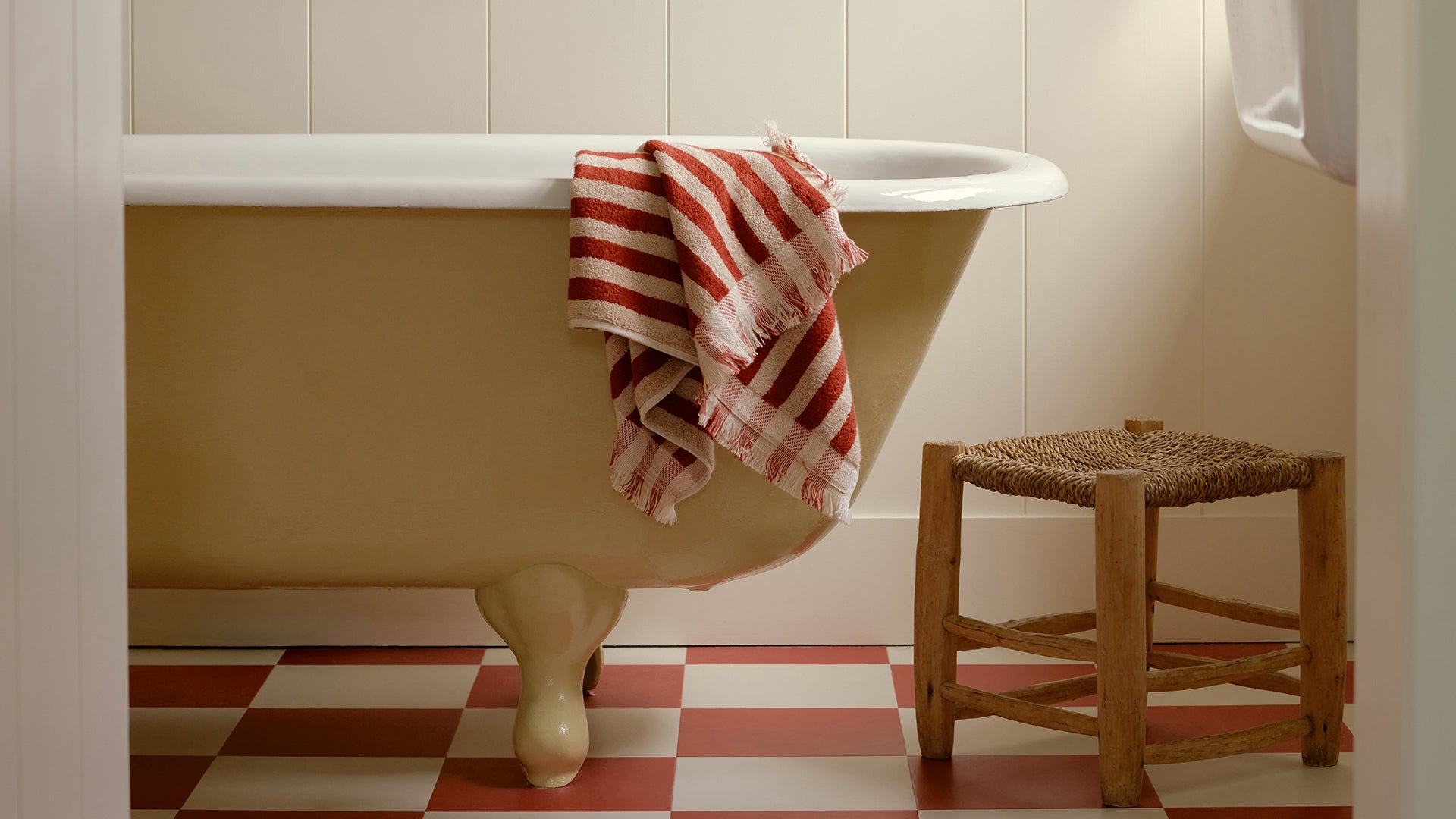 A vintage-style cream bathtub with claw feet in a bathroom, draped with a red and white striped towel, next to a small woven wood stool.