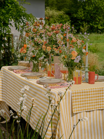 Outdoor table setting with a yellow and white checkered tablecloth, floral arrangements, and pink cups.