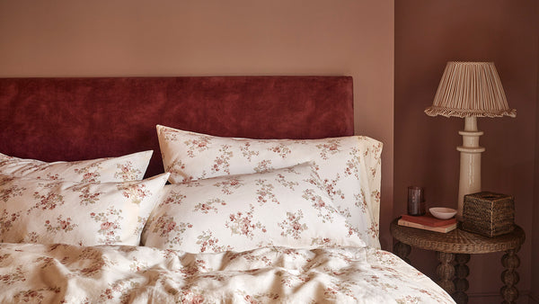A styled bedroom scene showing a bed with cream floral bedding, matching pillows, a dark red upholstered headboard, and a bedside table with a lamp against a warm brown wall.