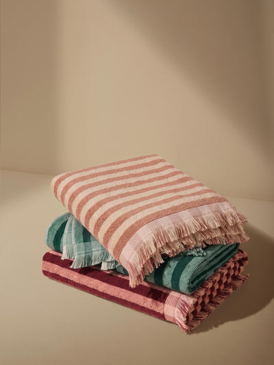 Three towels stacked on top of each other on a warm beige background. One is rhubarb and rose striped, one is pine green striped and one is sand shell striped. 