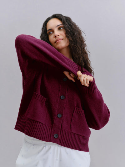 A woman wearing a maroon button-up knit cardigan with front pockets, posing against a solid light gray background.
