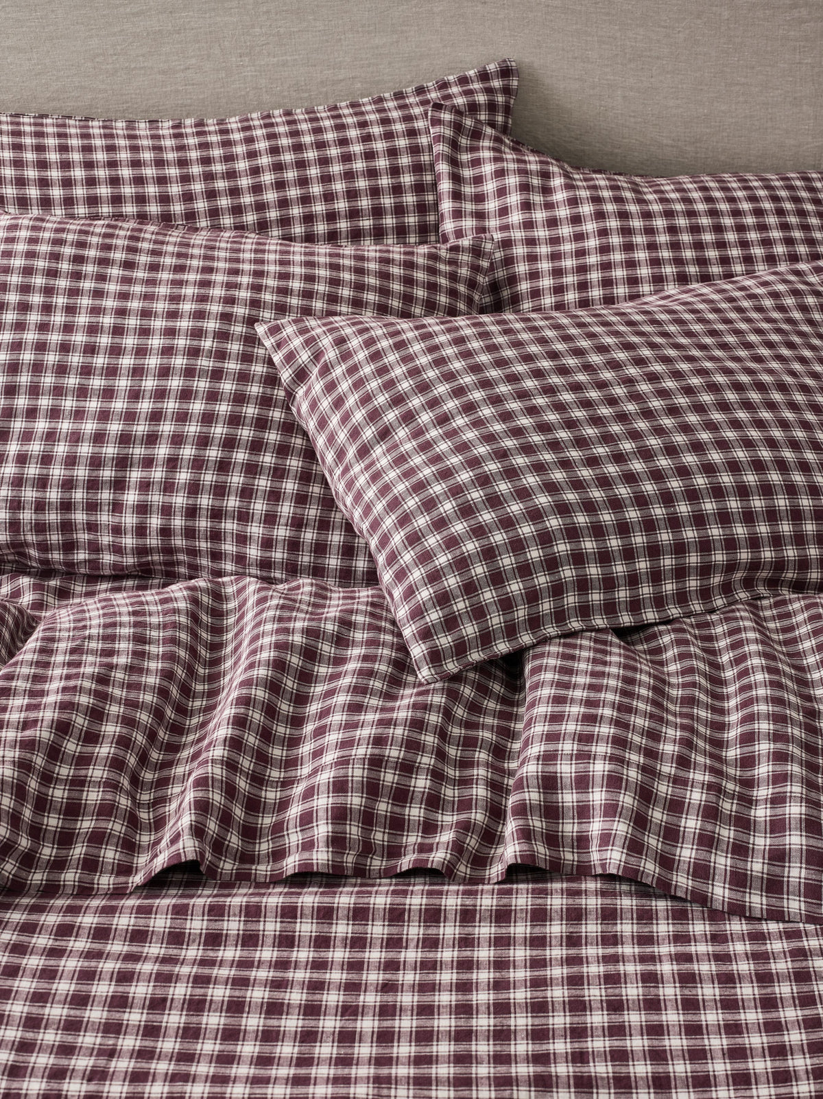 A close-up of a bed dressed in matching plaid bedding, including pillows and a sheet, in a dark burgundy and white checkered pattern against a neutral gray headboard.