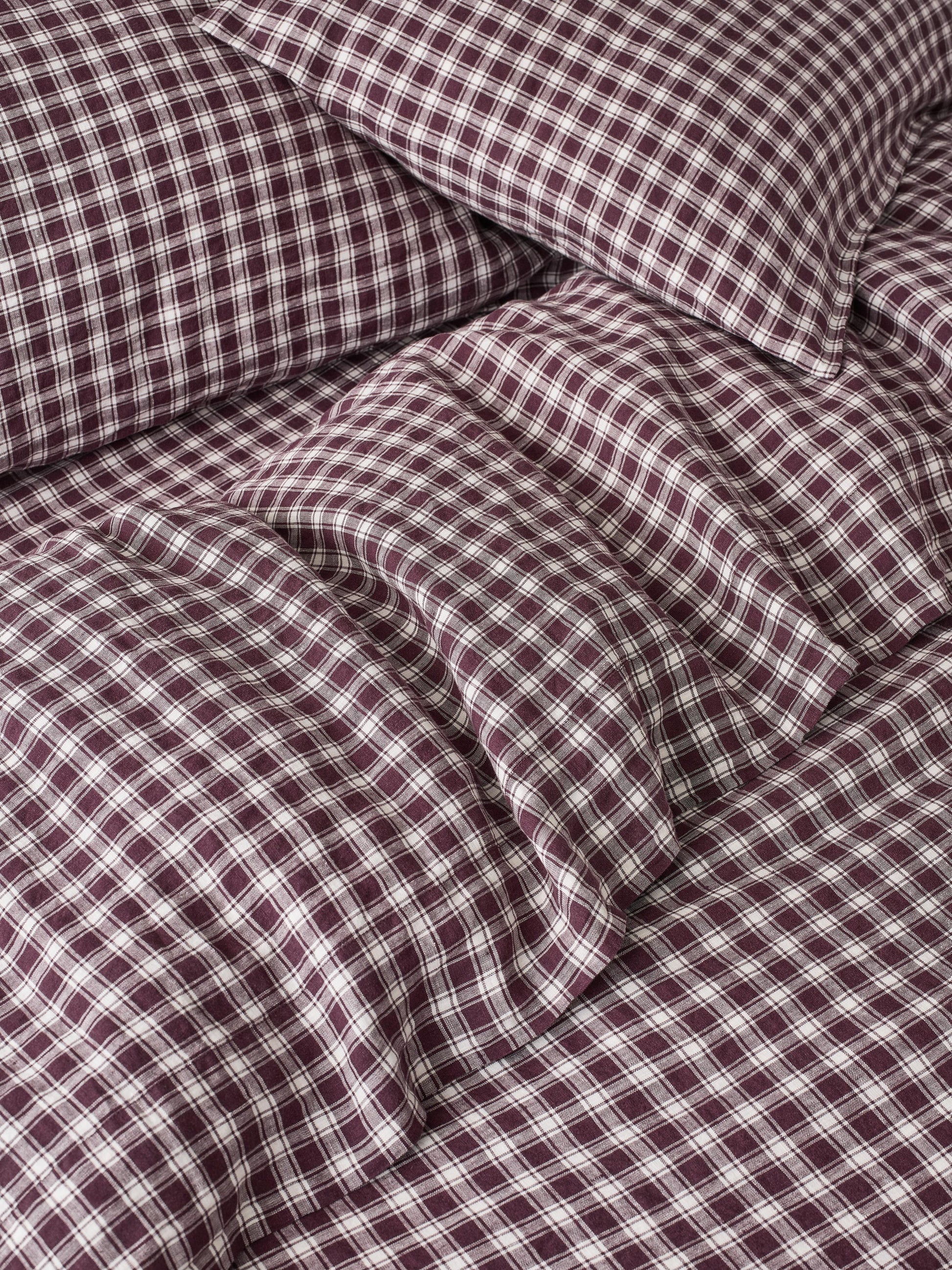A close-up of a bed dressed in matching plaid bedding, showing a duvet cover and pillowcase in a burgundy and white checkered pattern.