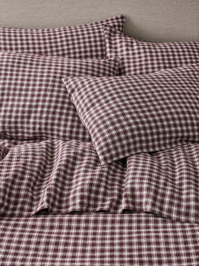 A close-up of a bed dressed in matching plaid bedding, including pillows and a sheet, in a dark burgundy and white checkered pattern against a neutral gray headboard.