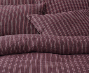 A close-up of a bed dressed in striped bedding, showing a duvet cover and matching pillowcases in a deep plum purple with thin vertical white stripes.