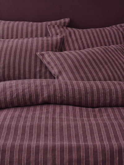 A close-up of a bed dressed in striped bedding, showing a duvet cover and matching pillowcases in a deep plum purple with thin vertical white stripes.