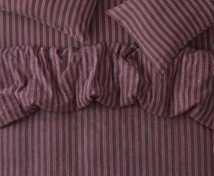 A close-up of a striped bedding set in a muted plum-purple color, featuring a ruffled duvet cover and two matching pillowcases.