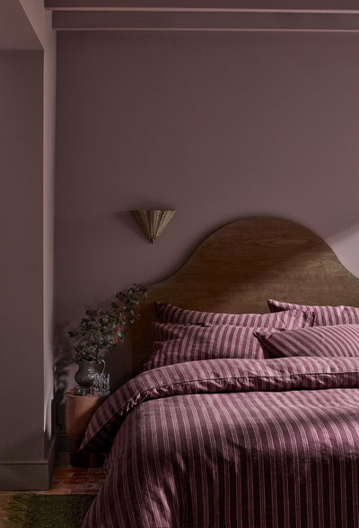 A cozy bedroom scene featuring a bed dressed in a dusty rose and burgundy striped duvet cover and matching pillow shams, set against a muted mauve-purple wall with a curved wooden headboard.