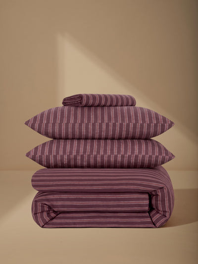 A stack of three folded bedding pieces, including two pillows and a folded sheet or duvet cover, in a dusty purple color with thin white vertical stripes, set against a warm beige background with a soft shadow.