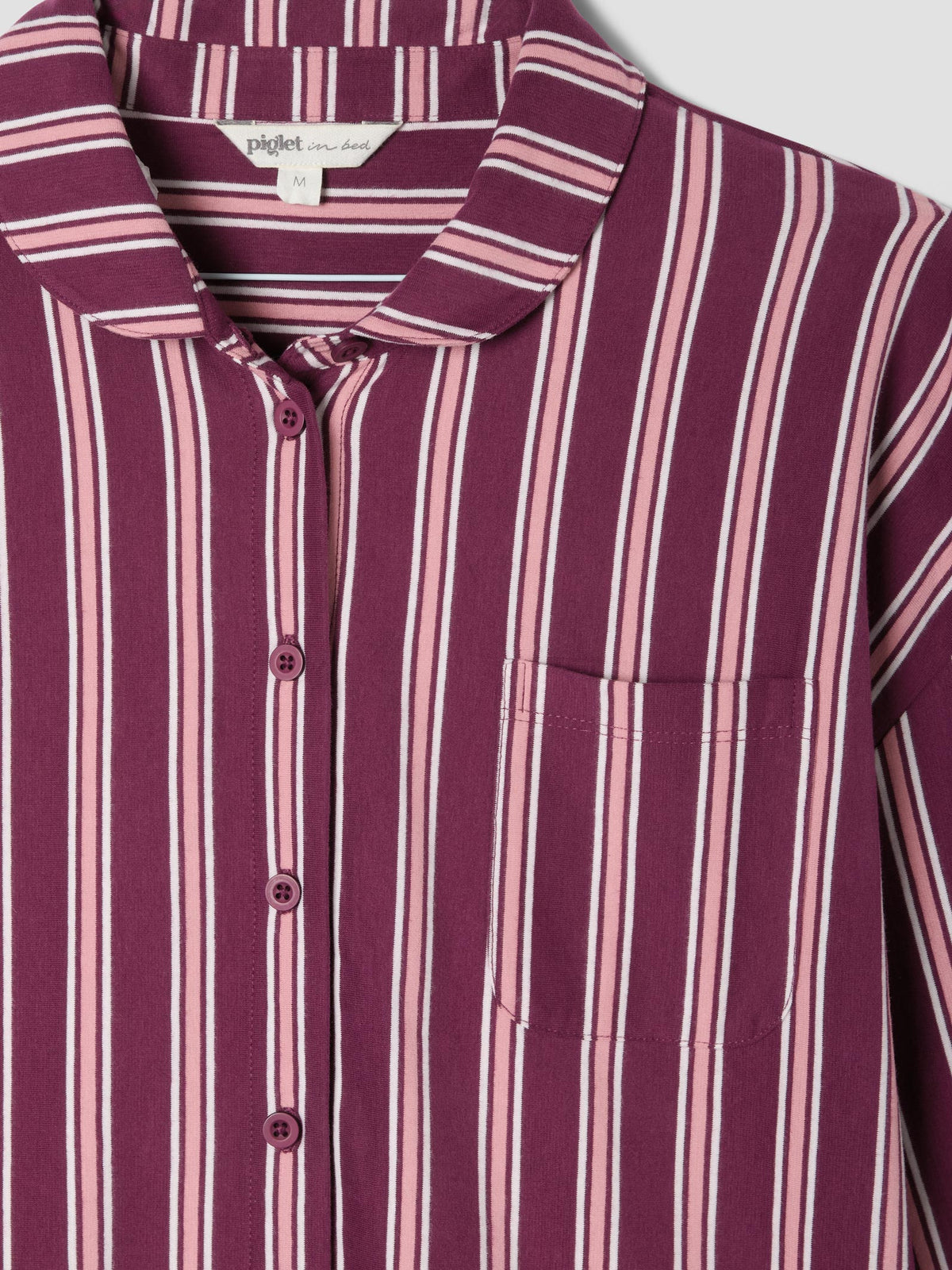 Close up of a long sleeved button up pajama top with pink and rhubarb red vertical stripes, and a peter pan collar on a light grey background.
