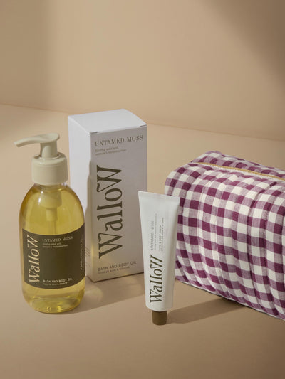 Wallow bath and body set including a pump bottle of Untamed Moss bath and body oil, a box of Untamed Moss, a tube of body cream, and a purple and white gingham zippered pouch on a beige background.
