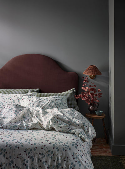 A styled bedroom scene featuring a bed with a dusty rose upholstered headboard, white bedding with a blue and brown floral pattern, and a small wooden bedside table holding a lamp with a brown shade and dried pinkish-red foliage against a dark gray wall.
