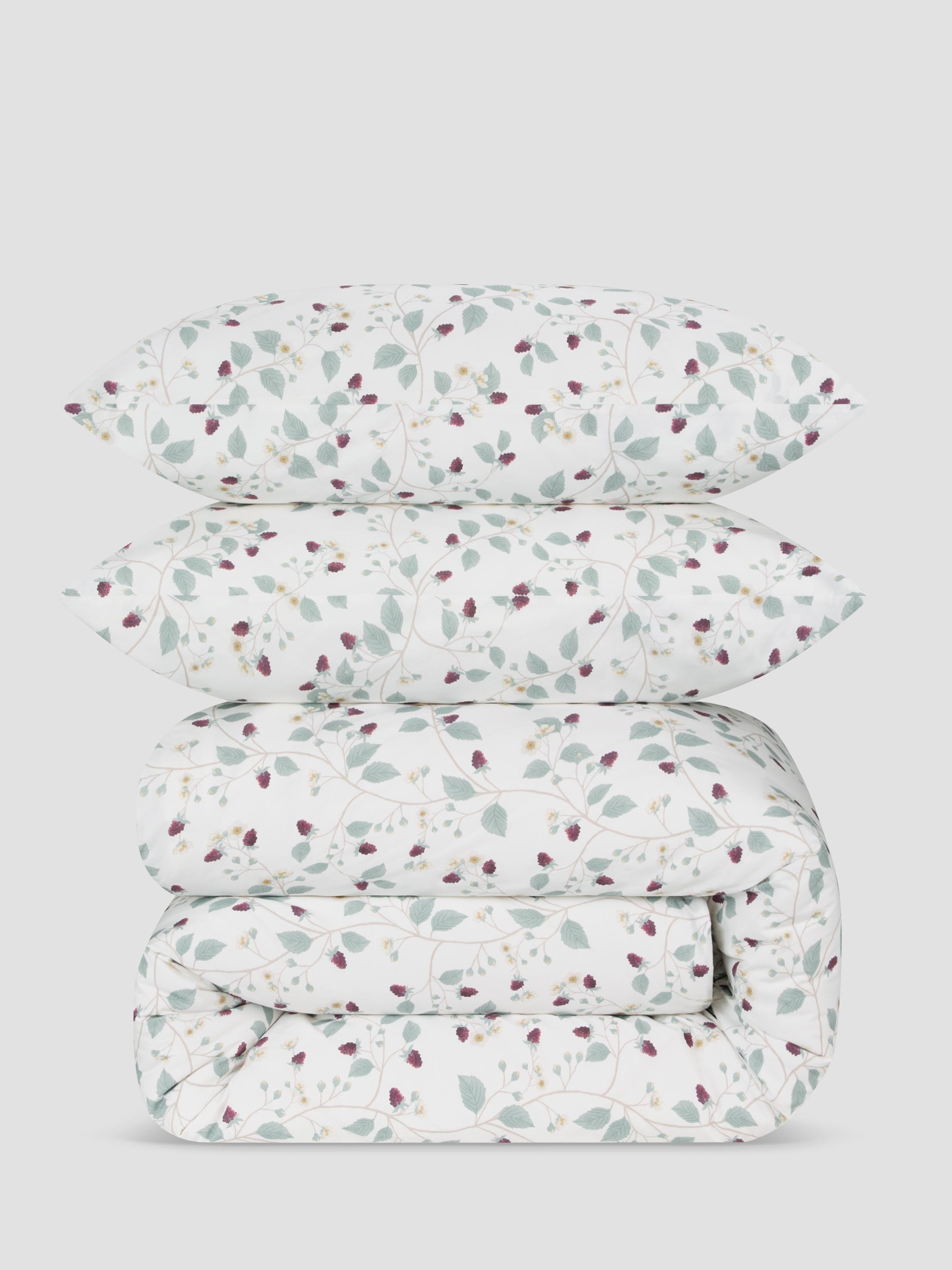 A stack of a folded duvet set, with matching pillowcases, featuring a delicate botanical pattern of sage green leaves and small burgundy berries on a white background, set against a solid light gray studio backdrop.