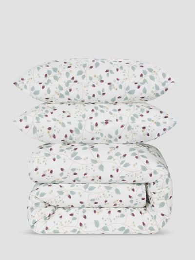 A stack of a folded duvet set, with matching pillowcases, featuring a delicate botanical pattern of sage green leaves and small burgundy berries on a white background, set against a solid light gray studio backdrop.