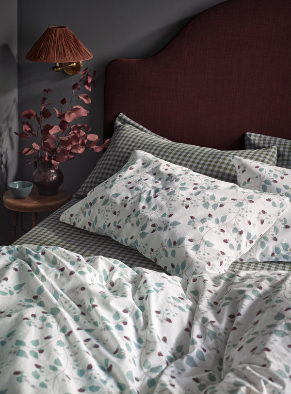 A styled bed in a dark bedroom setting featuring a white duvet cover and pillowcases with a delicate teal and burgundy botanical leaf pattern, layered over green-and-white gingham check sheets. A dark burgundy upholstered headboard, a bedside table with a lamp, and a vase of dried leaves complete the scene.