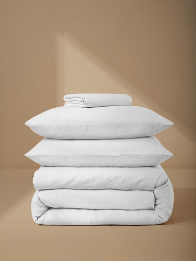 A stack of white bedding, including two pillows, a folded blanket, and a small folded towel, arranged on a warm beige background with soft diagonal shadows.