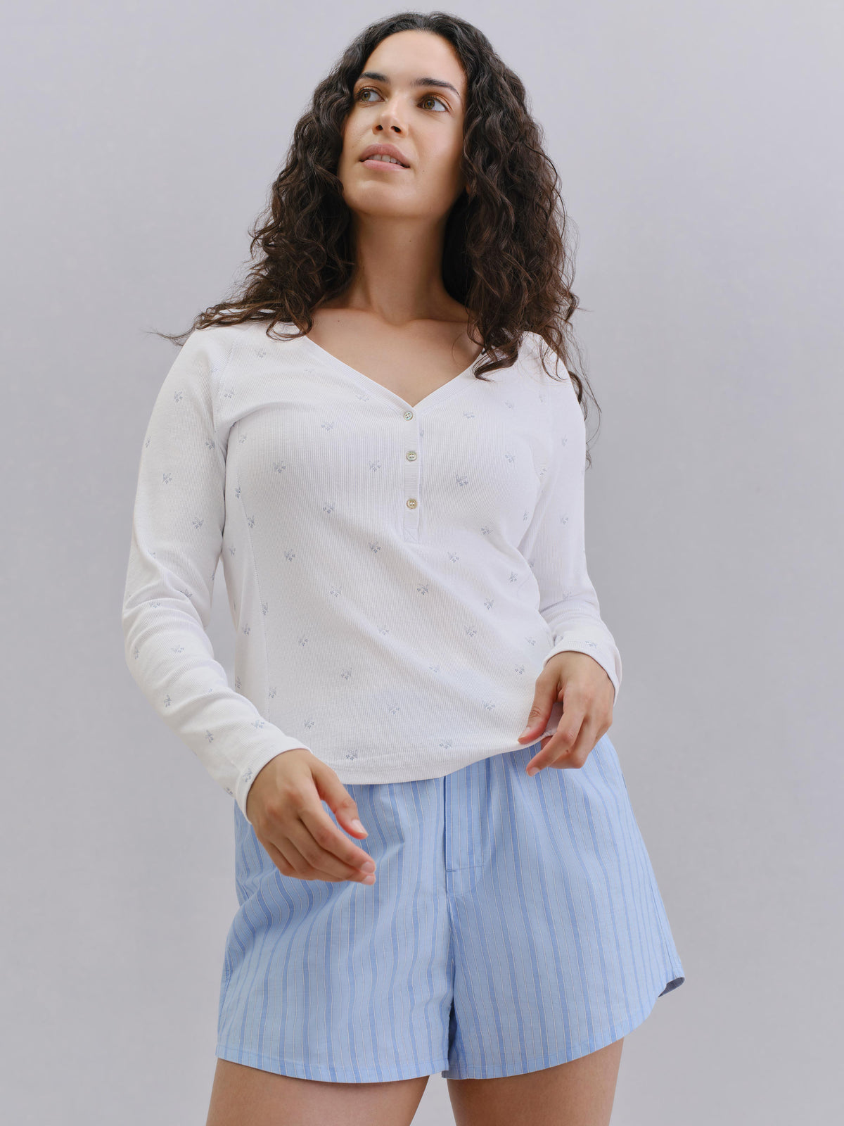 A woman modeling a white long-sleeve henley top with subtle floral prints and light blue pinstriped shorts against a plain gray studio background.
