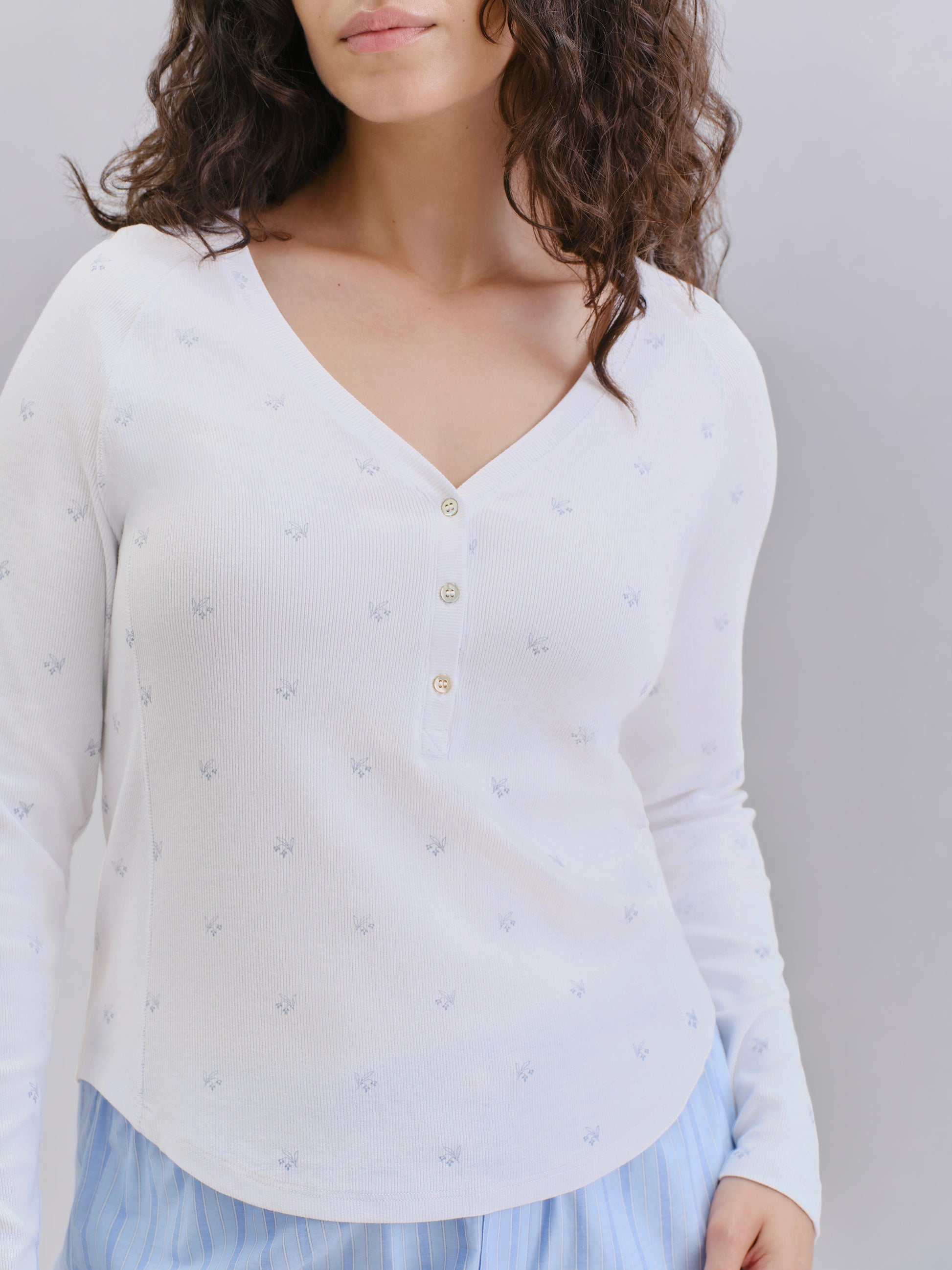 A woman wearing a white long-sleeve henley top with tiny blue floral prints and gold buttons, paired with a light blue skirt, against a neutral gray background.