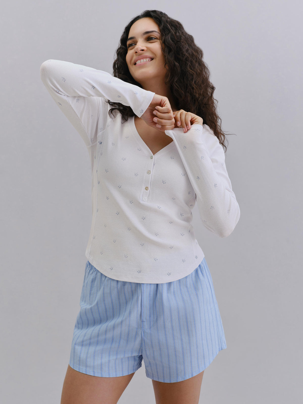 A woman modeling a white long-sleeve henley top with a subtle nautical pattern, paired with light blue and white vertically striped shorts, against a solid gray studio background.