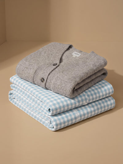 A folded gray knit cardigan layered on top of two folded light blue and white gingham shirts, arranged on a warm beige background.