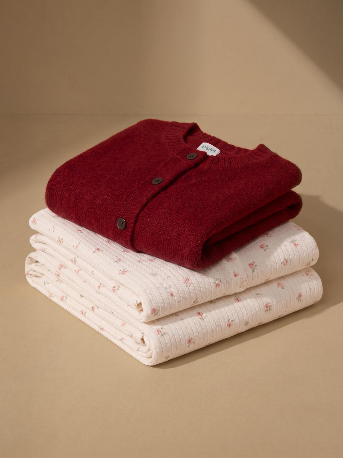 A folded burgundy knit cardigan layered on top of two folded cream pajama items with a small pink floral print, arranged on a warm beige background.