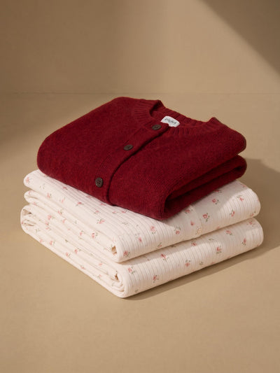 A folded burgundy knit cardigan layered on top of two folded cream pajama items with a small pink floral print, arranged on a warm beige background.