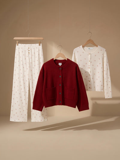 Three clothing items displayed on hangers against a warm beige studio backdrop: white patterned pajama pants, a burgundy cardigan, and a white cardigan with small pink floral prints.