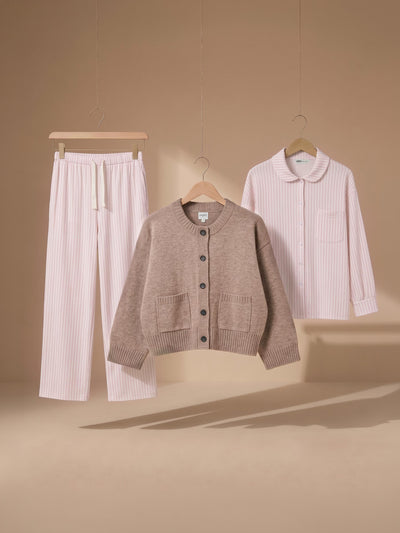A three-piece women's loungewear set hanging on wooden hangers against a warm beige background. The set includes light pink vertically striped pajama pants with a drawstring waist, a taupe knit cardigan with front pockets, and a matching light pink vertically striped long-sleeve top with a Peter Pan collar.