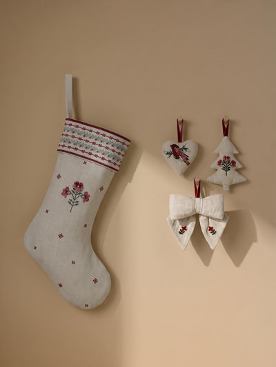A light beige linen Christmas stocking with embroidered pink flowers and a patterned cuff hangs on a warm beige wall, alongside matching fabric ornaments including a heart, a tree, and a bow.