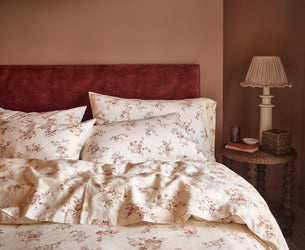 A cozy bedroom scene featuring a bed dressed in cream-colored floral bedding with a rust-red velvet headboard against a warm terracotta wall. A matching bedside table with a classic table lamp sits on the right.