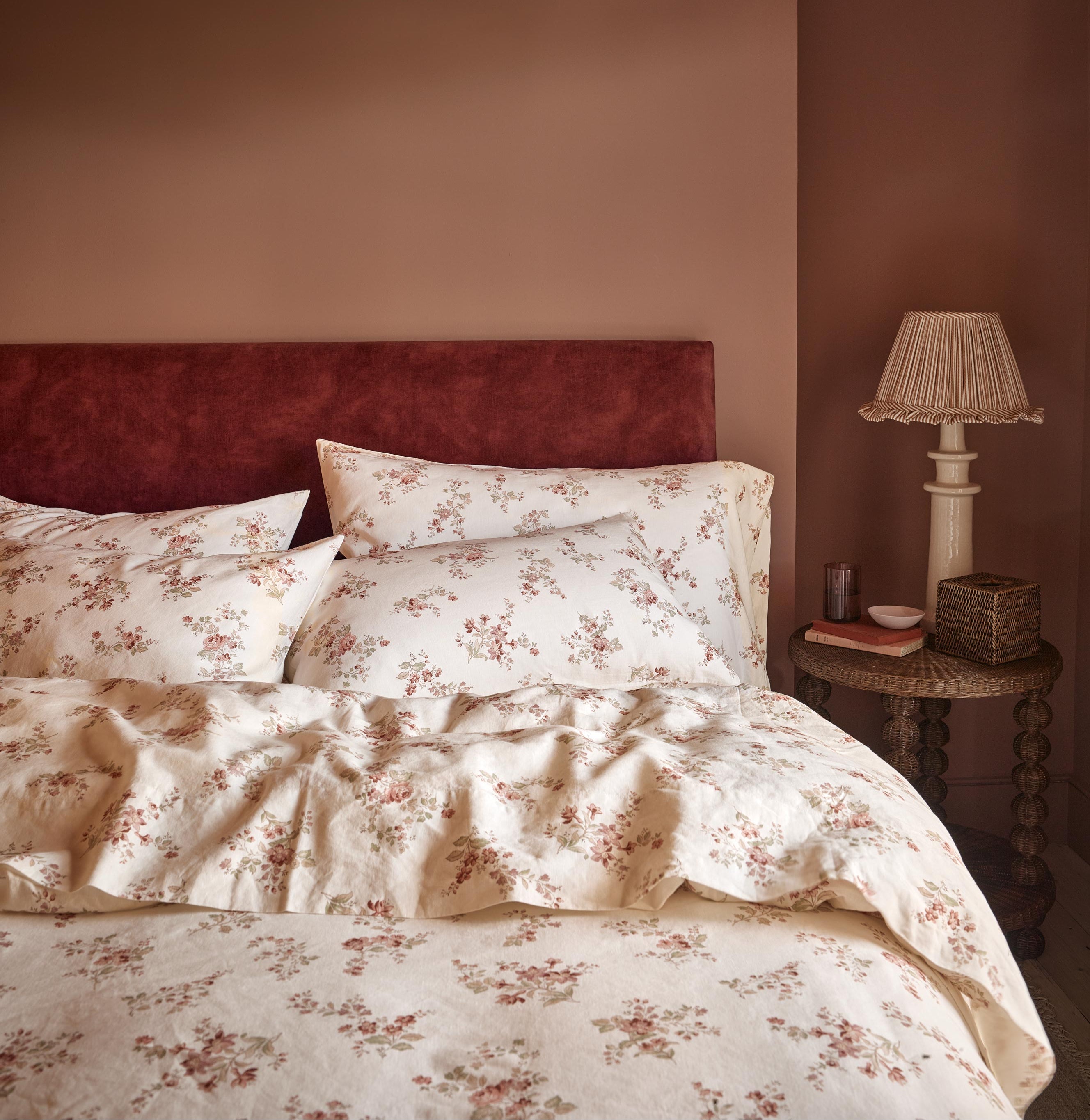 A cozy bedroom scene featuring a bed dressed in cream-colored floral bedding with a rust-red velvet headboard against a warm terracotta wall. A matching bedside table with a classic table lamp sits on the right.