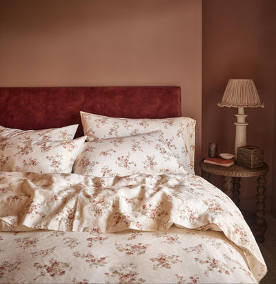 A cozy bedroom scene featuring a bed dressed in cream-colored floral bedding with a rust-red velvet headboard against a warm terracotta wall. A matching bedside table with a classic table lamp sits on the right.